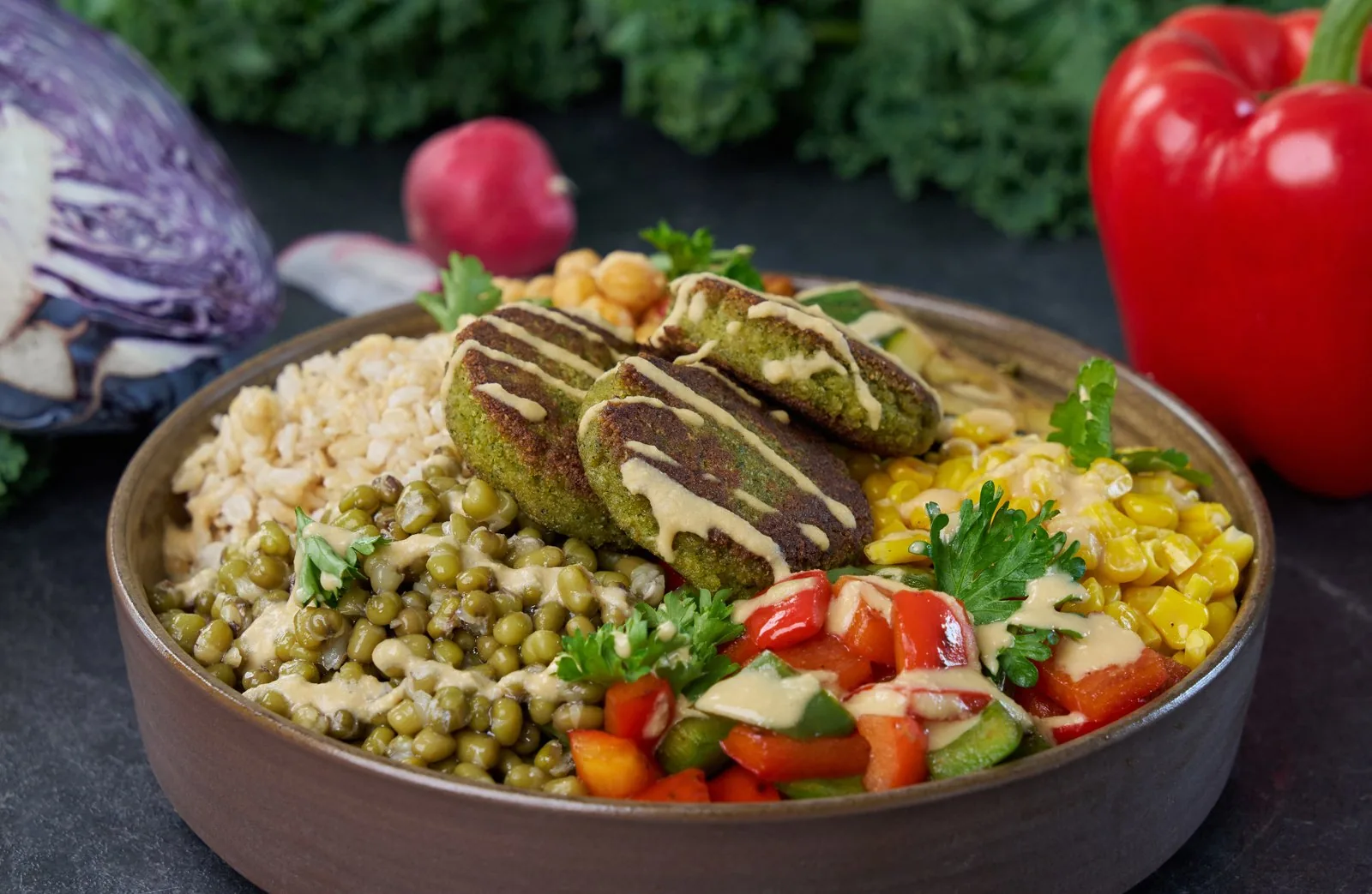 Professional food photography showing a wooden bowl with green falafel patties drizzled with tahini sauce, surrounded by white rice, green lentils, corn, fresh tomato and cucumber salad, red bell peppers, and fresh herbs